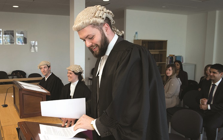 Students behind desks at a law moot, some wearing wigs, one smiling and looking at papers