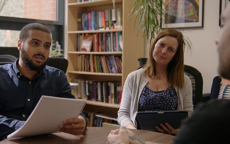 Two students with papers and a tablet, sitting behind a desk in a study, asking someone a question