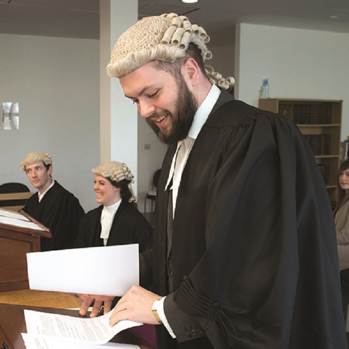 Students behind desks at a law moot, some wearing wigs, one smiling and looking at papers