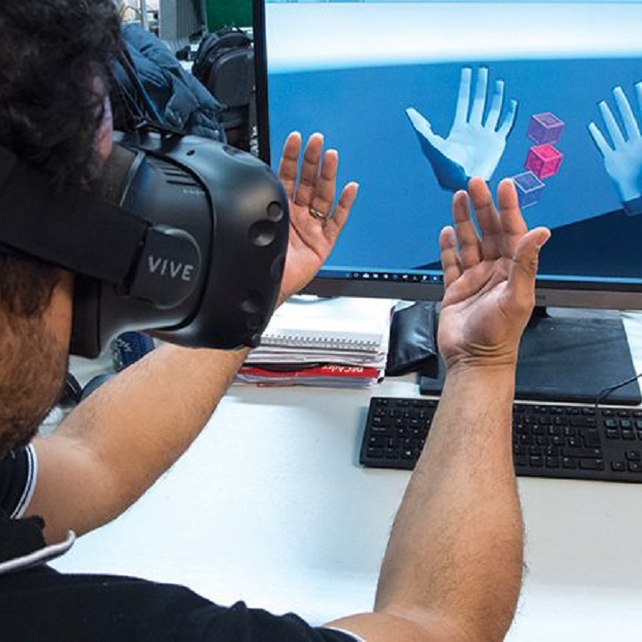 A student in a VR headset raising his hands while they display in virtual reality on a computer screen