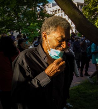 A black man in a black shirt stands aside from a crowd in the shade of a tree