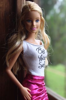 A Barbie doll stands by a window wearing a pink skirt and white top with the text Be Your Self written on it