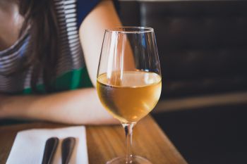 A close up shot of a glass of white wine with a woman wearing a blue and white stripey top in the background sitting at a restaurant table