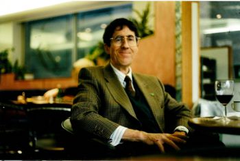 Dr (Jim) Perry James Browne seated by a table with a glass of red wine on it. He is smiling and wears glasses.