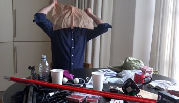 Vincent Gambini in a blue shirt with his head covered by a handkerchief standing behind a table covered in equipment for magic tricks