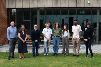 Members of the Pitch for the Planet judging panel and winning entrants stand in a line with the university campus as a backdrop