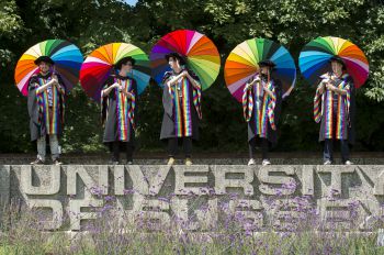 Five students stand atop the concrete University of Sussex sign wearing special rainbow gowns and holding rainbow umbrellas