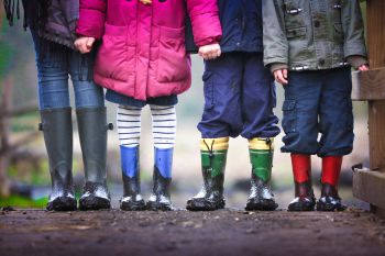 Four children stand facing the camera with muddy wellington boots. The image only pictures the children from the torso down.