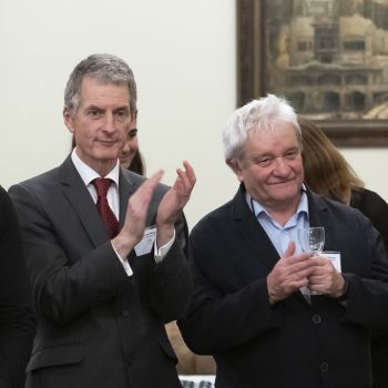 Two men dressed in suits standing side-by-side listening intently to a speech and applauding