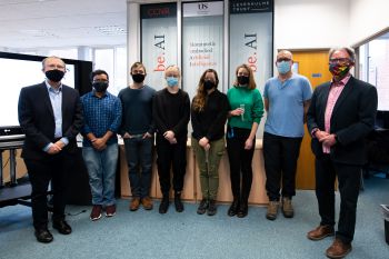 Two men in suits and wearing face masks stand either side of a line-up of six other people wearing more informal clothing and face masks