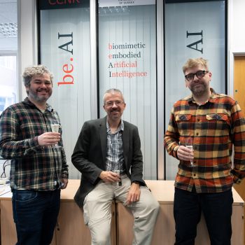 Two men standing either side of a third man sitting while all three hold champagne glassed