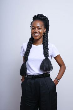 Fumani Mthembi stands with her hands in her pockets wearing a white shirt and black trousers with her hair in two plaits against a plain white backdrop