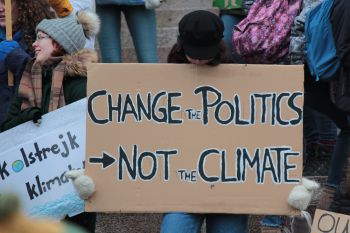 A person wearing a black hat and black coat stands in a protest holding up a cardboard placard