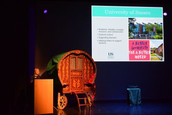 A woman speaks on stage with a screen and props around her