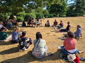 Postgraduate researchers sit in a circle outside on the ground listening to a speaker