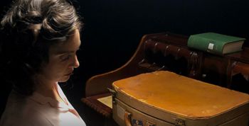 Woman in dramatic lighting, hair up, old briefcase sitting on a desk in front of her.