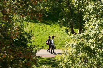 Two students walk on campus on a sunny day, they're walking past trees and grassy areas
