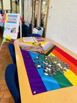 A progress pride rainbow flag laid out on a table with pronouns badges. In the background is a sign which says Sussex celebrates Trans Pride on a poster board.