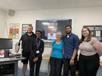 A group of four students and Margaret Kroto standing in a room with Graham Davis on the screen behind them