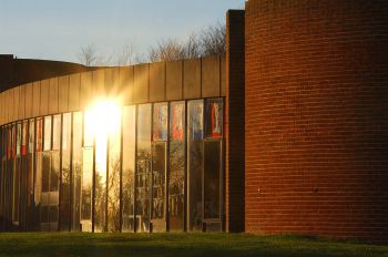 A photo of outside of the ACCA building on a clear blue sky sunny day. To the right third of the shot the curved wall of the building has orange bricks. The left two thirds of the shot feature the glass wall with the sun reflecting off it.