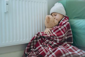 Photo of a child wrapped in a blanket wearing a woolly hat, seated on the floor and pressing against a radiator, holding a teddy bear.