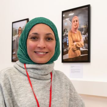 A Sussex colleague wearing a green headscarf in front of her picture which is part of the exhibition at the Library