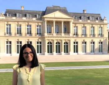 Picture of Yasemin Aslan, a woman with dark hair and a yellow top outside a building The Château de la Muette