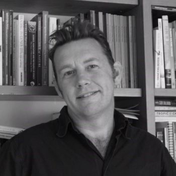 A black and white head and shoulders image of a smiling Professor Peter Newell standing in front of book shelves