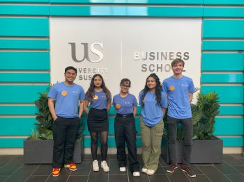 Five individuals stand in front of a wall with the Business School logo printed on it. The individuals are wearing blue t-shirts with a circular yellow badges on the chest.