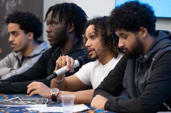 Four people sat in a row as a discussion panel, with one holding a microphone and talking