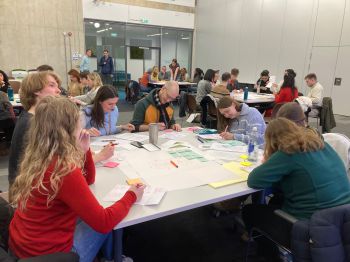 A room full of staff and students sit around tables, talking and writing with pen and paper. They are engaged in intense discussions, thinking deeply as they collaborate.