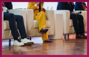 A panel of four people seated on white armchairs. The focus is on their lower bodies and legs. The person second from the left is wearing a bright yellow suit and purple high heels, while the others are dressed in dark suits with various footwear.