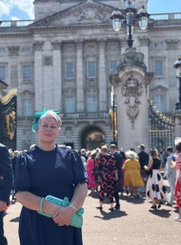 A smiling Lisa Holmes stands in front of the gates of Buckingham Palace. One can see other garden party guests arriving in the background.