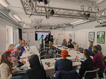 A group of people seated around U-shaped tables in a bright conference room, attending a workshop or meeting with a presentation screen at the front and three speakers standing nearby.