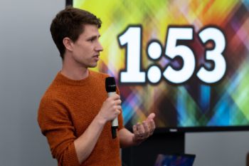 3MT presenter holding a microphone with a timer in the background