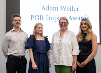 Four post-graduate researchers standing together and smiling with a screen behind them displaying the text