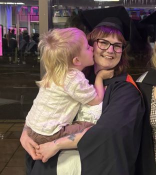 Rhiannon Williams is shown wearing her graduation robe grinning and holding her young son at her graduation ceremony in 2024