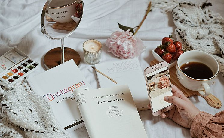A mobile phone taking a picture of a varied collection of items including strawberries, books about instagram, a mirror, a candle and watercolours